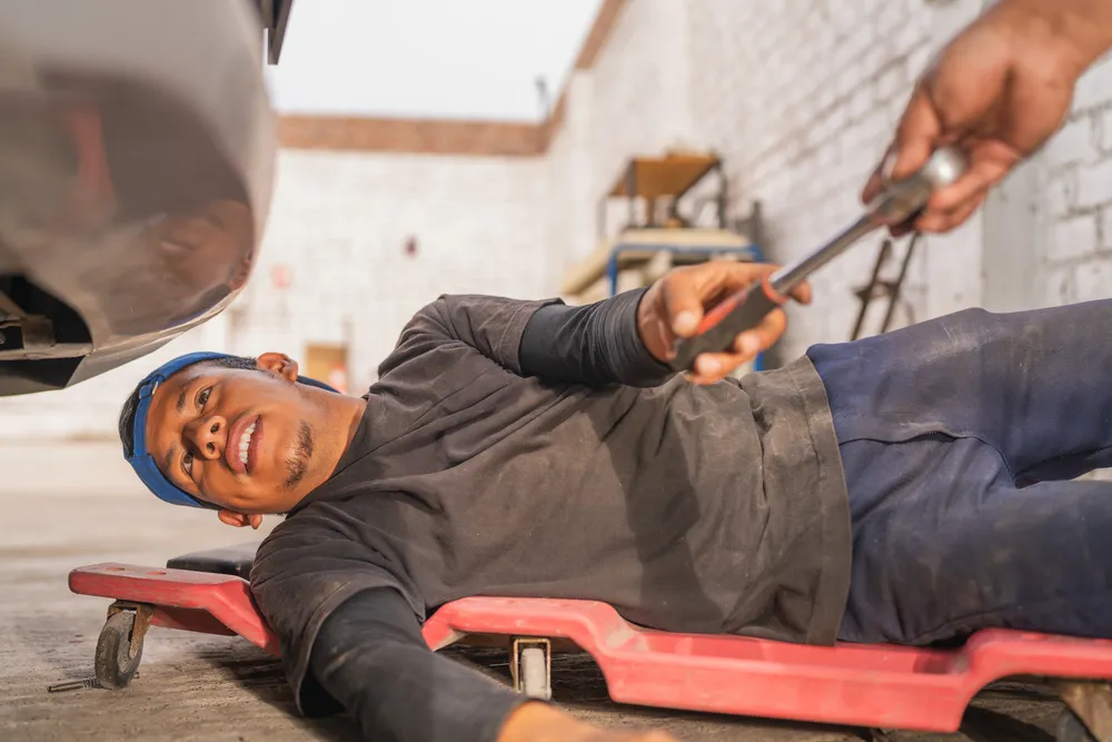 Skilled mechanic lying on the ground receiving a tool to check the undercarriage of a diesel truck as part of thorough diesel repair diagnostics and service