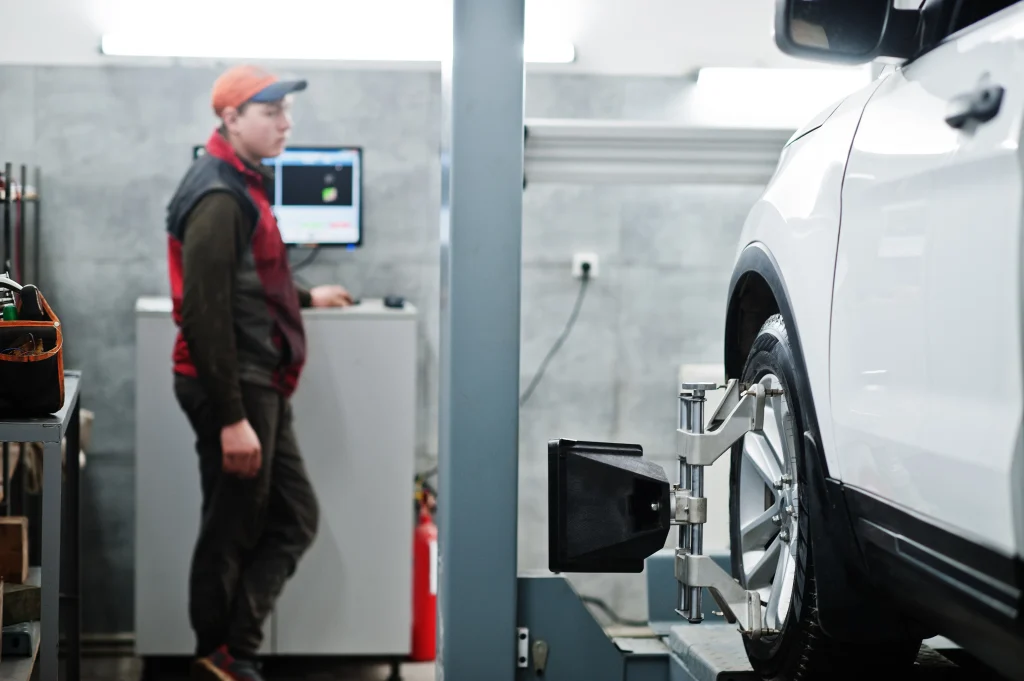 A mechanic in a cap and vest monitors the progress of wheel alignment calibrations on a car, with specialized equipment attached to the wheel.