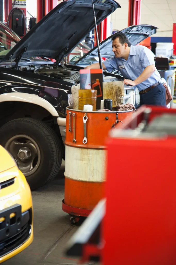 Mecánico apoyado en un coche abierto junto a un barril de herramientas durante una jornada de mantenimiento del auto.