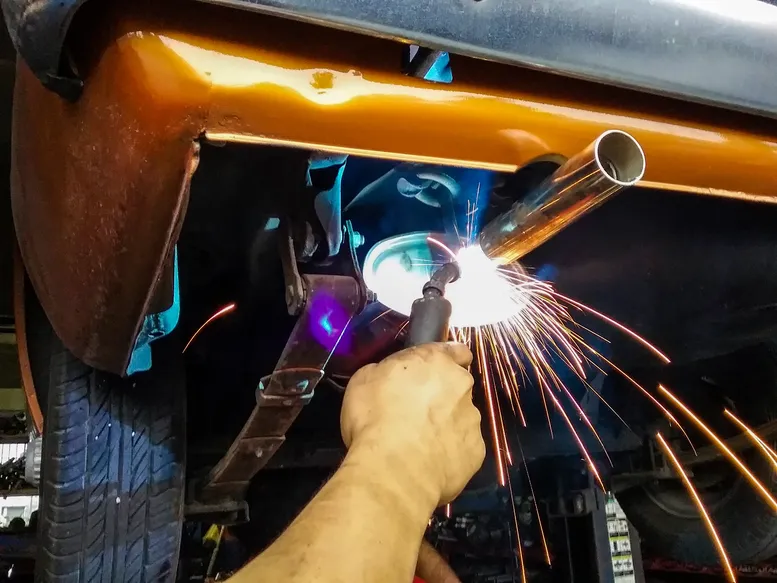 Welder applying a torch to a section of the exhaust pipe, creating bright sparks and an intense blue light.
