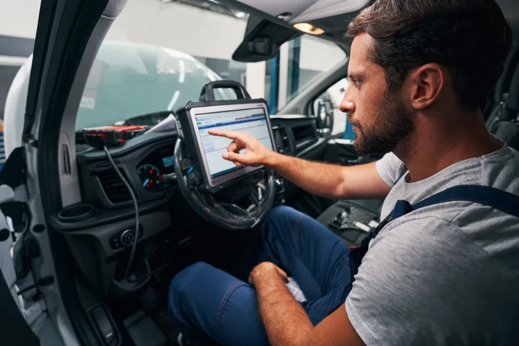 A mechanic sits in a car using a tablet to perform an engine diagnostic service.
