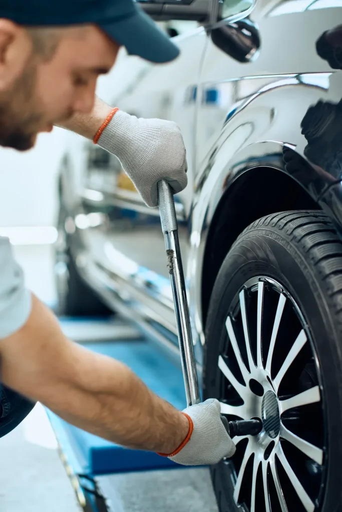 mechanic in a cap tightens the lug nuts on a car wheel with a lug wrench as a standard "car maintenance" task.